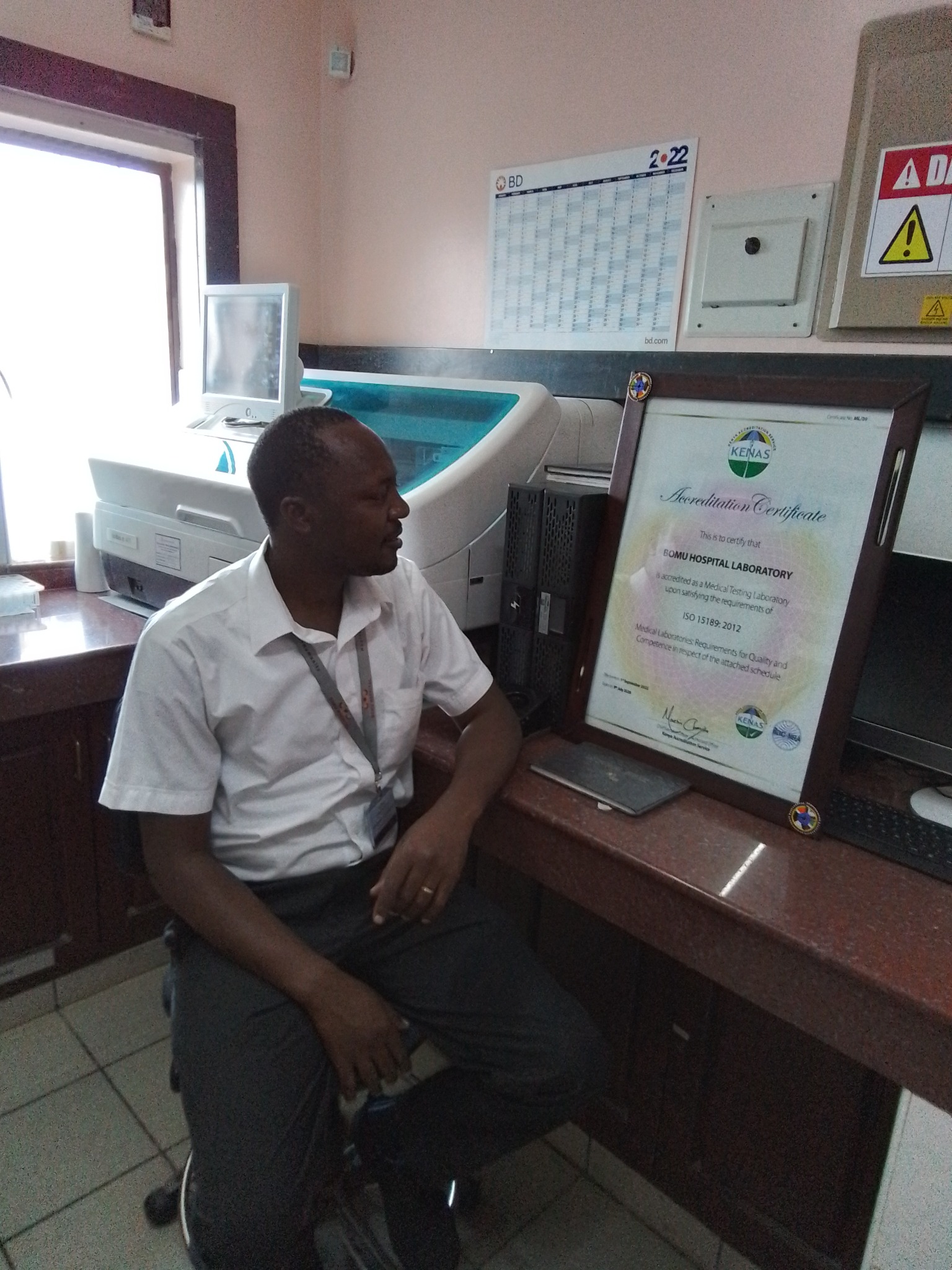 Japheth Mdenyo in a clinical laboratory beside a laboratory accreditation certificate